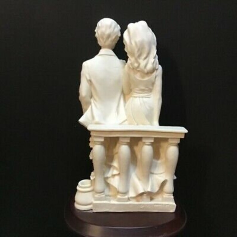 May include: White porcelain figurine of a couple standing on a balcony. The couple is dressed in formal attire and is holding hands. The balcony has a white railing and is decorated with flowers.