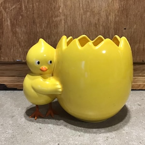 Puede incluir: Un macetero de cerámica amarillo en forma de pollito con una maceta de cerámica amarilla en forma de huevo. El pollito está sujetando la maceta de huevo con sus alas.
