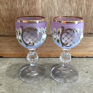 May include: Two purple glass goblets with gold rims and painted floral designs. The goblets have a clear glass stem and base.