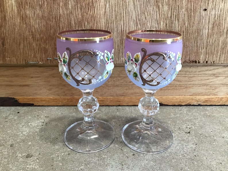May include: Two purple glass goblets with gold rims and painted floral designs. The goblets have a clear glass stem and base.