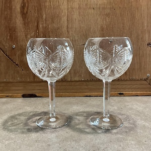 Op de afbeelding: Twee heldere kristallen wijnglazen met ingewikkelde patronen van sterren en geometrische vormen. De glazen hebben een afgeronde komvorm, een lange steel en een cirkelvormige voet. De achtergrond is een houten oppervlak.