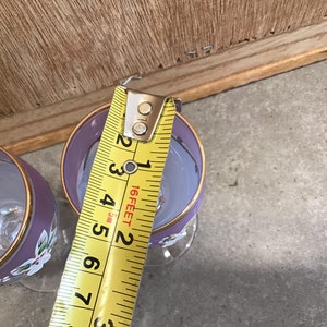 May include: Two purple glass wine glasses with gold trim and floral designs. A yellow measuring tape is used to measure the height of one of the glasses.
