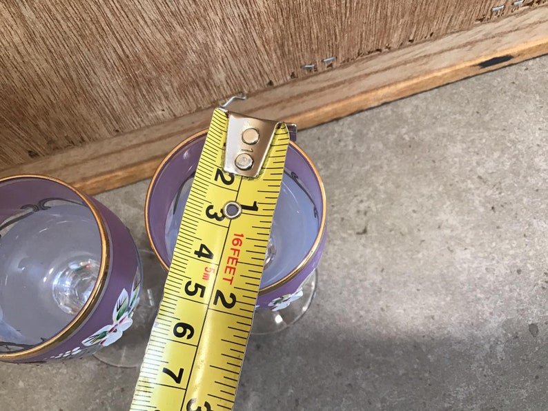 May include: Two purple glass wine glasses with gold trim and floral designs. A yellow measuring tape is used to measure the height of one of the glasses.