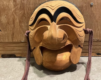 May include: Wooden mask with a smiling face, carved from light-colored wood. The mask features black accents above the eyes and a brown cord on each side. The mask has a large nose and open mouth.