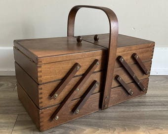 May include: A vintage wooden sewing box with a hinged lid and a wooden handle. The box has multiple compartments and angled wooden supports. The wood has a warm, brown tone, and the box appears to be well-crafted and sturdy.