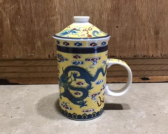 Puede incluir: Una taza de té de cerámica amarilla con tapa, que presenta un diseño de dragón azul y detalles intrincados. La taza tiene un asa y una tapa con un pomo.