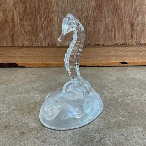 May include: A clear glass sculpture of a seahorse on a base with a textured surface.
