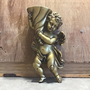 May include: A gold cherub figurine holding a trumpet. The cherub has wings and is wearing a flowing robe. The figurine is made of resin and is suitable for indoor use.