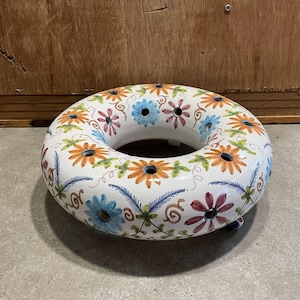 Peut inclure: Un anneau en céramique blanche avec un motif floral dans des tons d'orange, bleu, rose et vert. L'anneau a un trou au centre et est décoré de petits trous autour du bord.