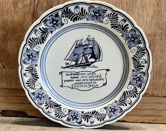 May include: A white ceramic plate with a blue floral border and a central image of a sailboat. The plate features a Dutch inscription in a banner below the ship. The plate has a scalloped edge.