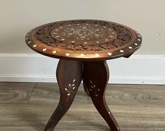 May include: A small, round, dark brown wooden table with intricate carved floral designs on the tabletop. The table has three curved legs and decorative inlaid accents. The table is approximately 18 inches in diameter.