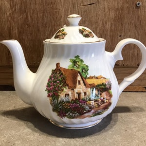 Könnte beinhalten: Weiße Porzellan-Teekanne mit einem Cottage-Motiv auf der Seite. Das Cottage ist braun mit einem Strohdach und von Blumen und Grün umgeben. Die Teekanne hat einen goldenen Rand um den Deckel und die Basis.