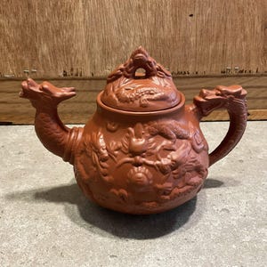 May include: A terracotta-colored teapot with intricate dragon relief carvings. The teapot features a dragon-shaped spout and handle, and a lid with a dragon finial. The body of the teapot is adorned with detailed dragon imagery.