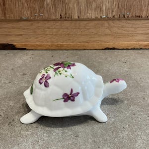 May include: A white ceramic turtle figurine with a purple and green floral design. The turtle is sitting on a gray surface.