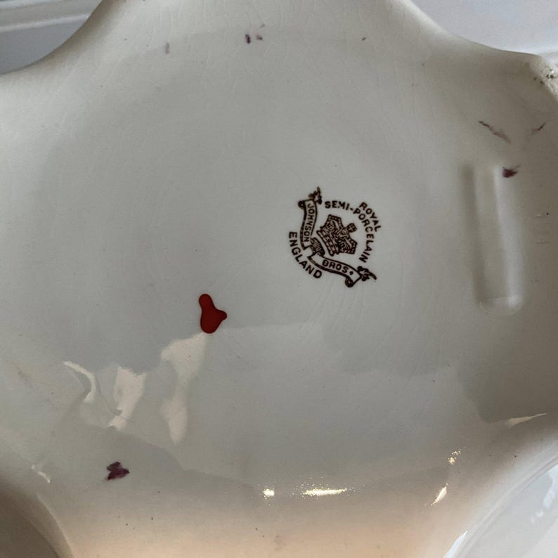 May include: Close-up of a white porcelain dish with a star-like shape. The dish has a brown and red emblem that reads "Johnson Bros. England Royal Semi-Porcelain". There are small red marks on the surface of the dish.