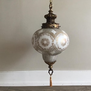 Może przedstawiać: Biała i złota lampa wisząca w kształcie kuli z ozdobnym frędzlem. Lampa ma mosiężne wykończenie i skomplikowane detale.