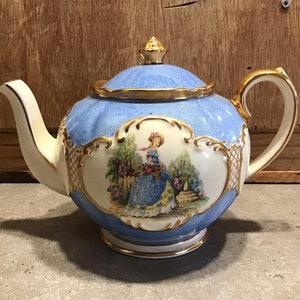 May include: A vintage porcelain teapot with a light blue body, gold trim, and a lid with a gold finial. The teapot features a painted scene of a woman in a garden. The handle and spout are cream-colored.