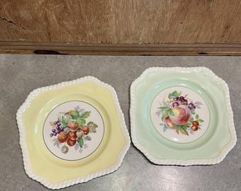Puede incluir: Dos platos decorativos con bordes festoneados. Uno es amarillo con un diseño de fresas y uvas. El otro es verde claro con un diseño de melocotón y uvas. Ambos platos tienen un borde blanco en forma de cuerda.