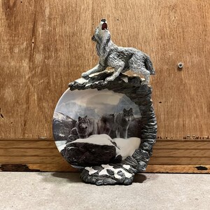 May include: A ceramic plate with a wolf howling at the moon design. The plate is surrounded by a sculpted rock formation with a wolf figurine on top. The plate features a snowy scene with two wolves in the background.