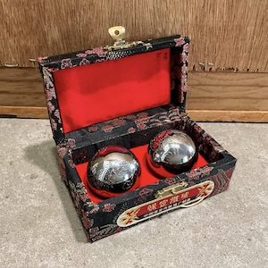 Può includere: Una scatola nera con interno rosso contiene due sfere metalliche argentate lucide. La scatola è decorata con un motivo floreale rosso e oro. La scatola ha una chiusura dorata. Testo cinese sulla scatola.