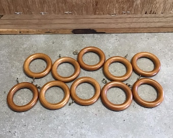 Könnte beinhalten: Zehn Gardinenringe aus Holz mit goldenen Haken. Die Ringe sind hellbraun und haben eine glatte Oberfläche.