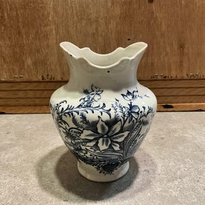 Könnte beinhalten: Eine weiße Keramikvase mit einem ausgestellten, gewellten Rand. Die Vase weist ein detailliertes blaues Blumenmuster auf, einschließlich großer Blüten und Blätter. Die Vase hat einen abgerundeten Körper und einen kleinen Sockel.
