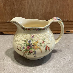 Può includere: Brocca in ceramica color crema con disegni floreali. La brocca presenta un corpo arrotondato, un manico curvo e un beccuccio. Il corpo è decorato con motivi floreali in rilievo e fiori dipinti a mano in vari colori, tra cui rosso, viola e giallo.