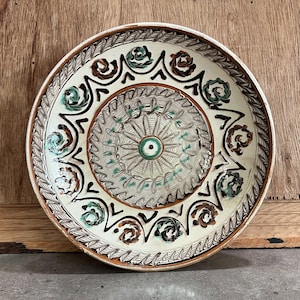 May include: A decorative ceramic plate with a white base, featuring intricate hand-painted designs. The plate has a brown rim and a central pattern with green and brown floral motifs. The outer edge has a repeating leaf pattern.