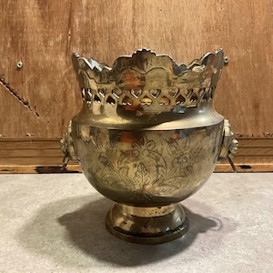 Puede incluir: Una jardinera de latón vintage con intrincados grabados florales y de hojas. La jardinera presenta un borde decorativo con recortes en forma de corazón y asas de cabeza de león. La base es un diseño de pedestal.