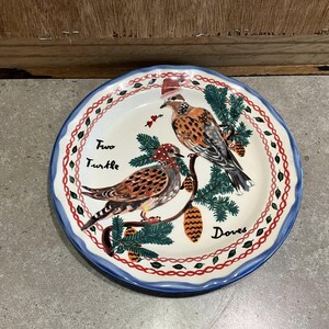 Może przedstawiać: Okrągły ceramiczny talerz z niebieską obw&oacute;dką i dekoracyjną czerwoną i białą obw&oacute;dką. Talerz przedstawia malowany wz&oacute;r dw&oacute;ch gołębi siedzących na gałęzi z igłami sosnowymi i szyszkami. Na talerzu widnieje napis "Two Turtle Doves".