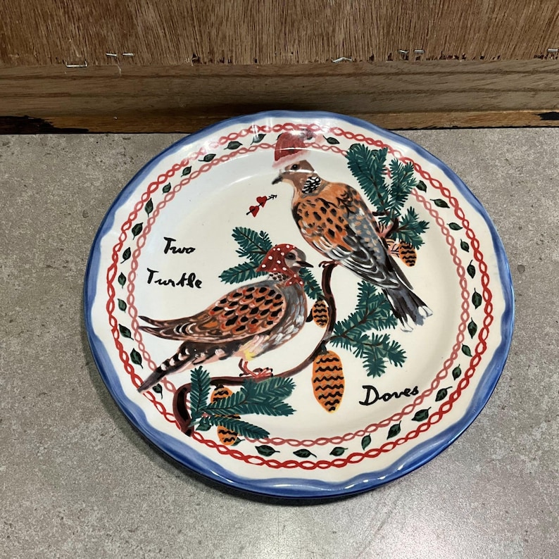 Może przedstawiać: Okrągły ceramiczny talerz z niebieską obw&oacute;dką i dekoracyjną czerwoną i białą obw&oacute;dką. Talerz przedstawia malowany wz&oacute;r dw&oacute;ch gołębi siedzących na gałęzi z igłami sosnowymi i szyszkami. Na talerzu widnieje napis "Two Turtle Doves".