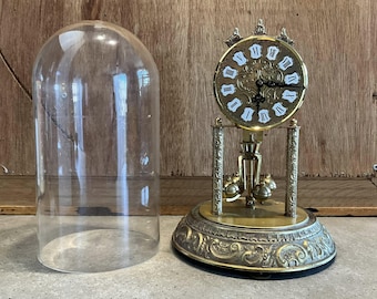 Op de afbeelding: Een antieke messing klok met een witte wijzerplaat en zwarte Romeinse cijfers, op een sierlijke basis. Een heldere glazen koepel staat links. De klok heeft een slinger en decoratieve elementen.