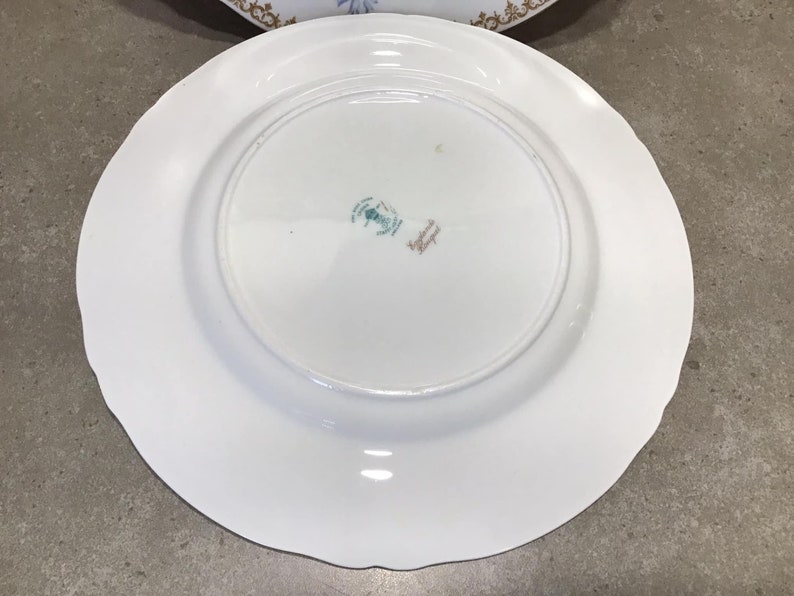 May include: White ceramic plate with a scalloped edge. The back of the plate has a green and gold mark that reads "Made in England".