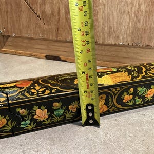 May include: A black decorative box with floral and figure designs, measured with a yellow tape measure. The tape measure shows a length of approximately 6 inches.