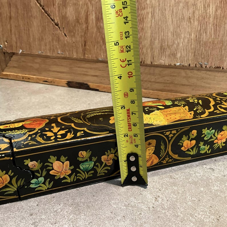 May include: A black decorative box with floral and figure designs, measured with a yellow tape measure. The tape measure shows a length of approximately 6 inches.