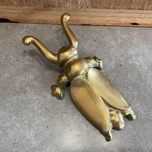 Może przedstawiać: Złoty metalowy podnośnik do butów w kształcie stylizowanego owada. Podnośnik ma długie, wąskie ciało z zakrzywioną głową i dwoma uniesionymi ramionami.