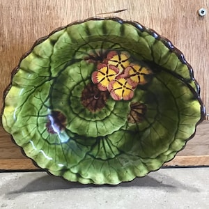 Può includere: Un piatto in ceramica verde con bordo ondulato, che presenta un design a foglia stilizzato e fiori dipinti in tonalità di giallo, rosa e marrone.