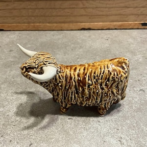 Puede incluir: Figura de cerámica de una vaca de las Tierras Altas en tonos marrón y beige. La figura tiene un pelaje lanudo, cuernos blancos y una cara detallada. La vaca está de pie sobre cuatro patas pequeñas, y el diseño general es rústico y encantador.