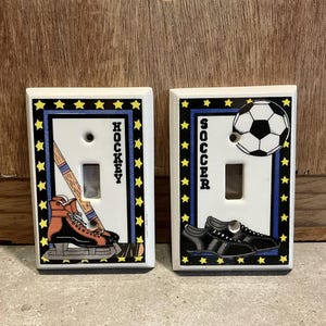 May include: Two white ceramic light switch covers with a blue and yellow star border. The left cover features a hockey stick, puck, and ice skate with the word "HOCKEY" written vertically. The right cover features a soccer ball with the word "SOCCER" written vertically.