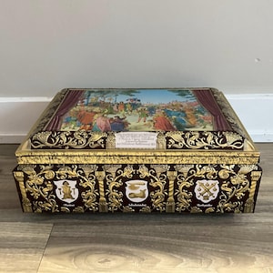 May include: A rectangular burgundy and gold tin box with a detailed painting on the lid depicting a historical scene. The box features ornate gold scrollwork and crests on the sides, with text visible on the lid.