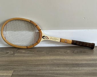 Puede incluir: Raqueta de tenis Dunlop vintage con marco de madera y mango blanco y marrón. La raqueta presenta el nombre "Marty Riessen" y un retrato en el mango. La empuñadura está envuelta en cuero marrón.