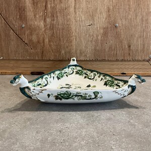 May include: A white ceramic serving dish with a boat-like shape, featuring green floral patterns and dragon head accents on each end. The dish has a small handle on the top and gold trim.