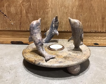 May include: A stone candle holder featuring three carved dolphins in a circle around a small candle holder. The base is a round stone with a textured surface.
