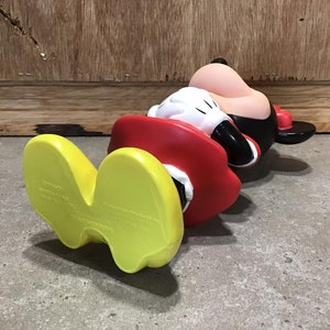 May include: A Mickey Mouse figurine in a lying down position. The figure is wearing a red jacket, black shorts, and a black hat. The feet are yellow, and the gloves are white. The background is a wooden wall and a concrete floor.