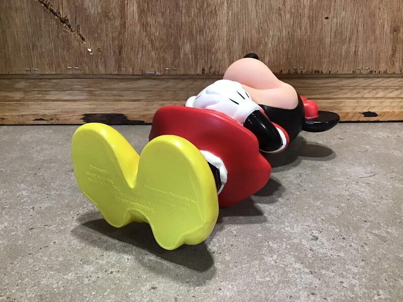 May include: A Mickey Mouse figurine in a lying down position. The figure is wearing a red jacket, black shorts, and a black hat. The feet are yellow, and the gloves are white. The background is a wooden wall and a concrete floor.
