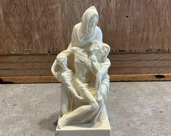 May include: White ceramic sculpture depicting the Pietà, a scene of Mary holding the body of Jesus after his crucifixion. The sculpture is detailed and shows the sorrow and grief of the figures.