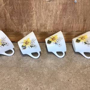 Pode incluir: Quatro ch&aacute;venas de ch&aacute; de cer&acirc;mica branca com desenhos florais amarelos e brancos. As ch&aacute;venas est&atilde;o dispostas em uma fila, com as al&ccedil;as viradas para a direita.