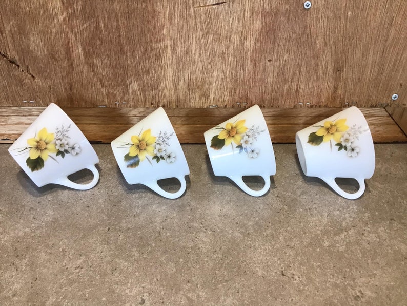 Pode incluir: Quatro ch&aacute;venas de ch&aacute; de cer&acirc;mica branca com desenhos florais amarelos e brancos. As ch&aacute;venas est&atilde;o dispostas em uma fila, com as al&ccedil;as viradas para a direita.