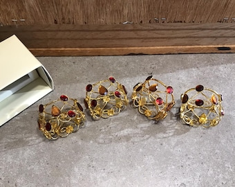 May include: Four gold-toned napkin rings with red and orange gem accents. The rings are intricately designed with a floral pattern.