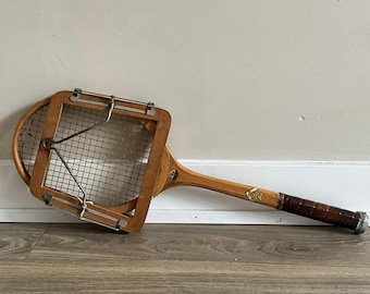 Könnte beinhalten: Ein antiker Tennisschläger aus Holz mit Ledergriff und einem Drahtgeflecht. Der Schläger liegt auf einem Holzboden.
